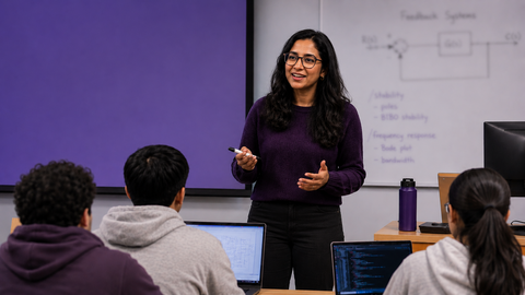 AI-generated image of a female teaching assistant standing at the front of a modern engineering classroom speaking to students seated with laptops open. A blurred whiteboard with technical diagrams is visible in the background, creating a collaborative graduate learning environment.