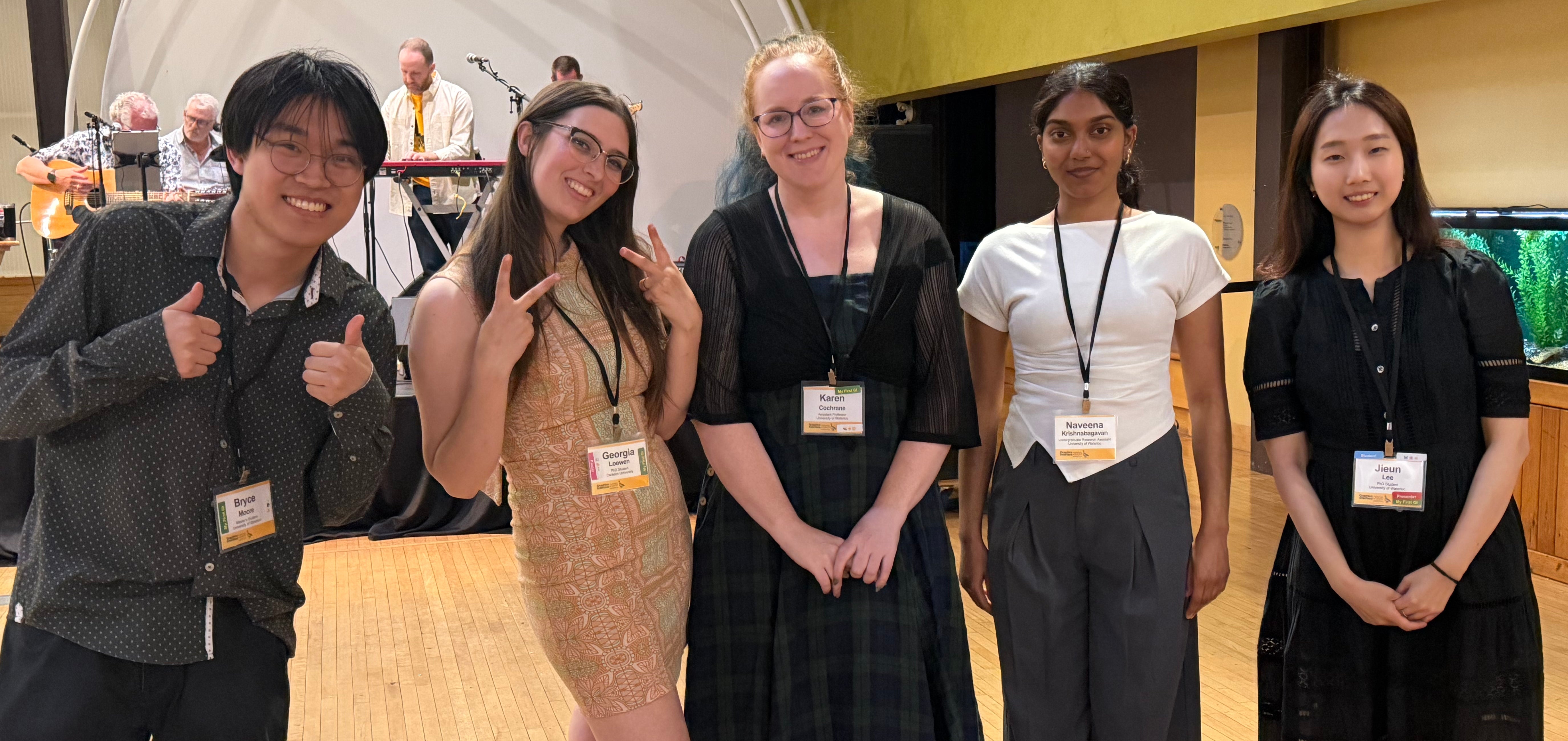 Five members of the Embodied Computing Lab pose together at an event, smiling at the camera. They are standing in a row in front of a stage with musicians and instruments in the background.