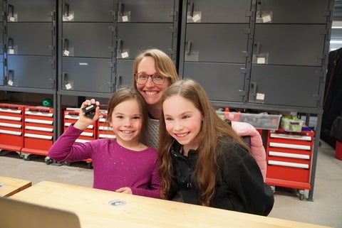 A mother and her two daughters smiling at the camera, one daughter is holding up a micro:bit