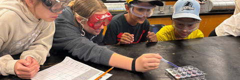4 campers with lab googles on looking at an science experiment, while one camper pipettes a substance into a tray