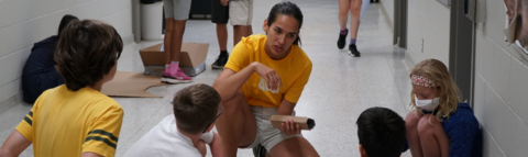 Camp leader holding cardboard, talking to four campers who are building and sitting on the floor in a hallway