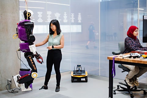 A woman standing in front of a humanoid robot, while another sits in front of a computer. 