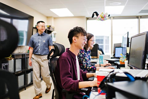 Two students working on computers, while a smiling professor looks over their shoulders