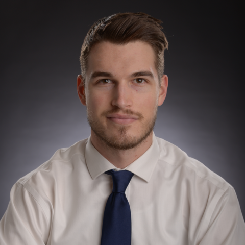 Photo of Alex wearing a white shirt and blue tie, smiling at the camera