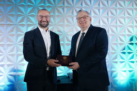Two men stand in front of a blue wall sharing a plaque.