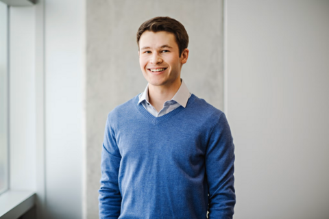 A man in a collared shirt smiles at the camera in front of a white wall