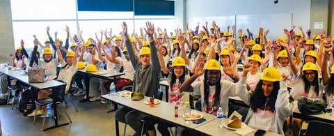 A classroom full of students with their arms up and wearing yellow hard hats.