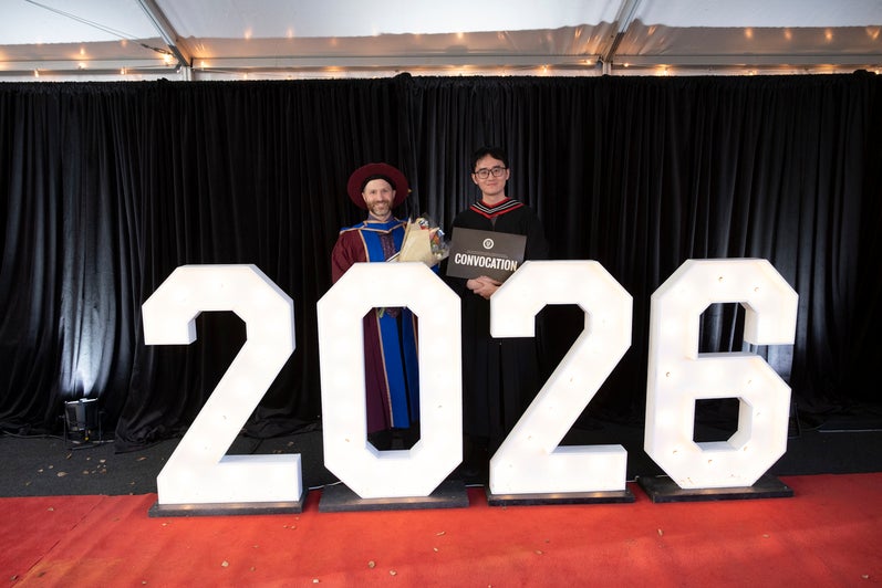 Two people stand behind the 2026 sign outside of convocation