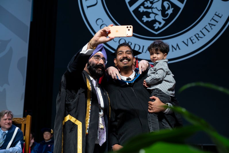 Two adults and a child smile as a selfie is being taken while the middle man is being hooded
