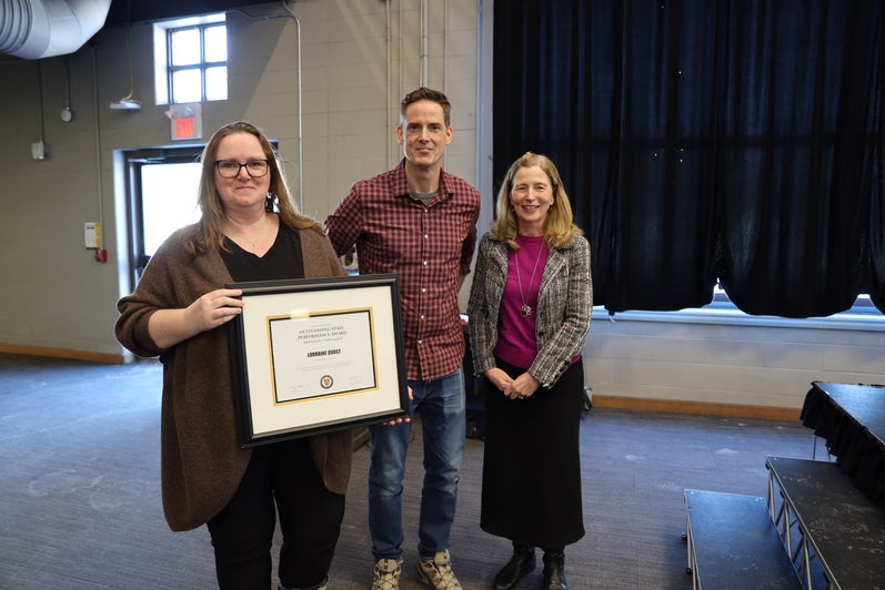 A woman, a man and another woman pose beside the stage with a certificate.