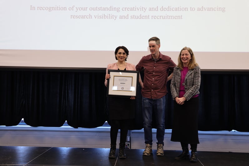 A woman, a man and another woman pose on stage with a certificate.
