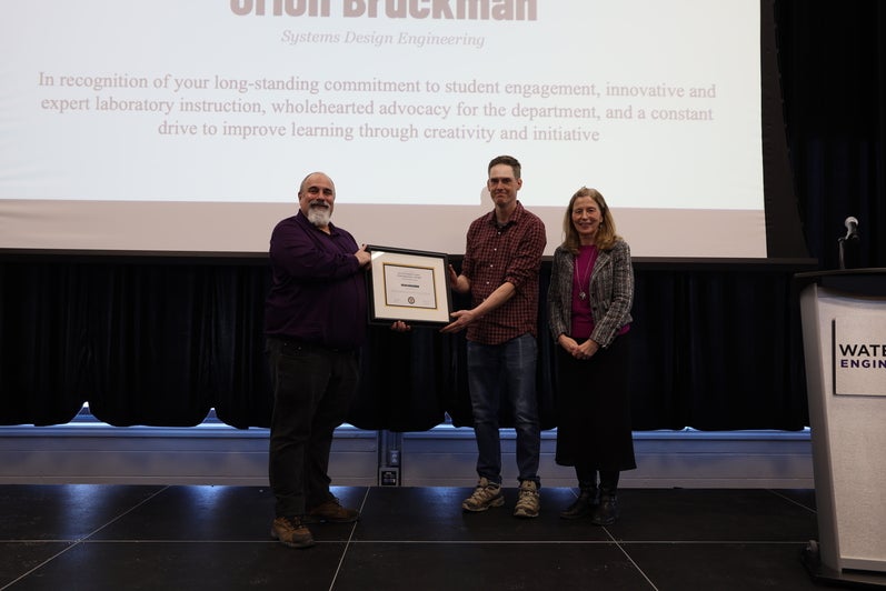 Two men and one woman pose on stage with a certificate