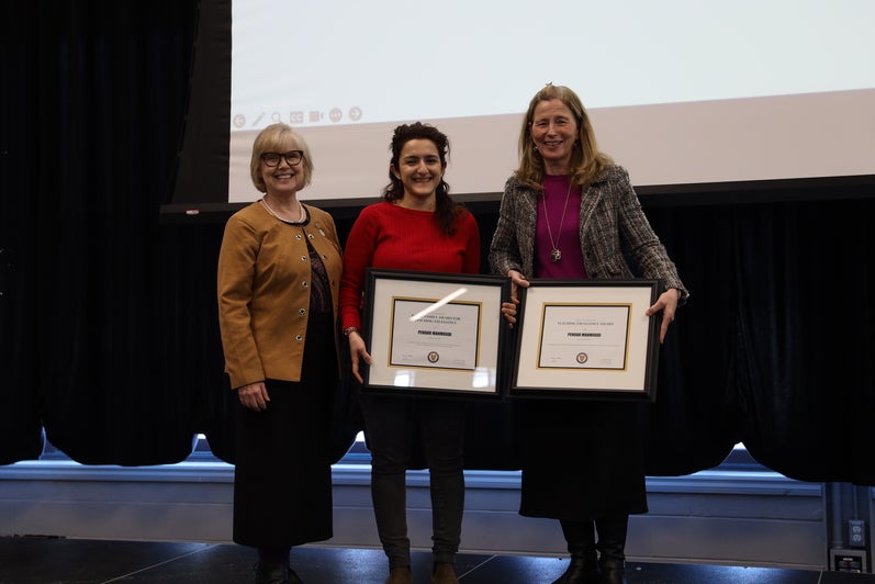 Three women pose on stage with two framed certificates between them