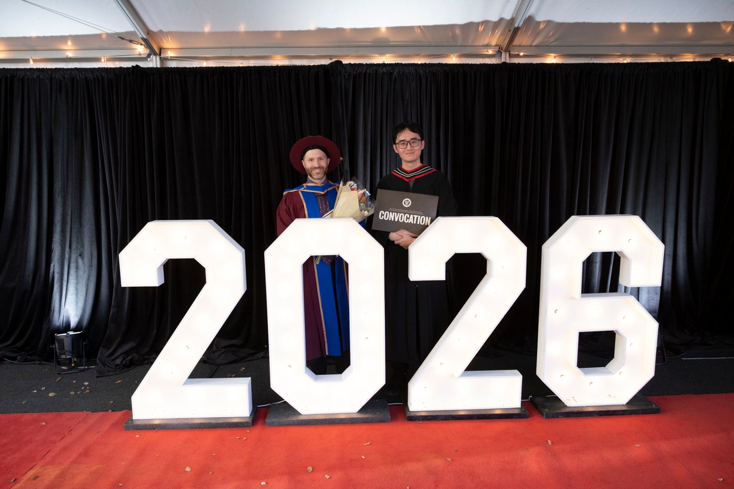 Two people stand behind the 2026 sign outside of convocation