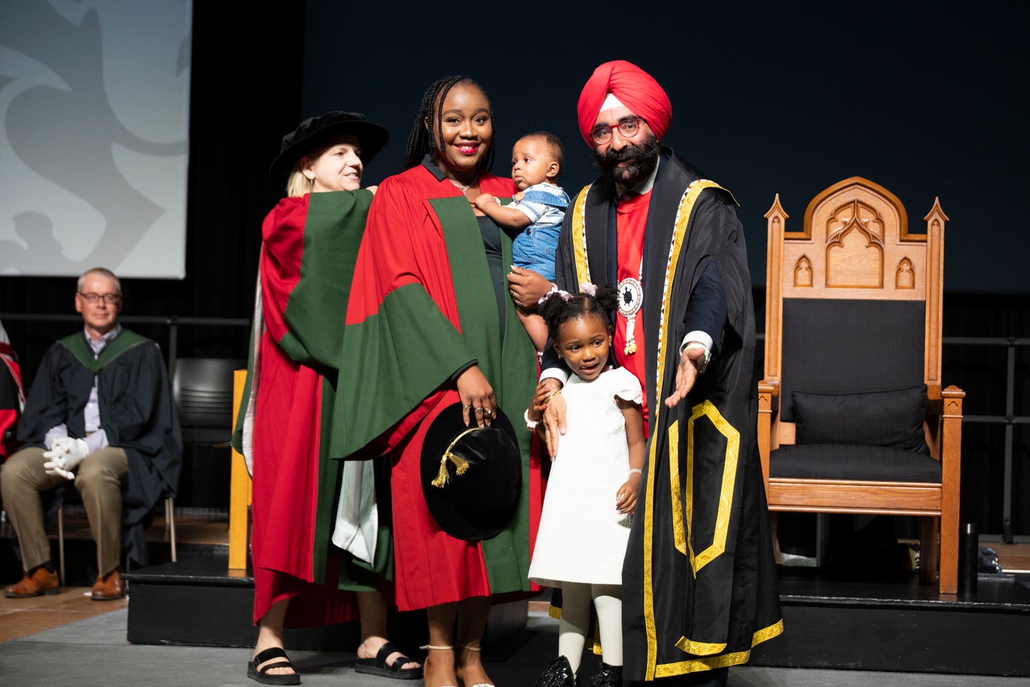 A woman is receiving her graduate hood, holding her baby and the hand of her young daughter, on stage at convocation