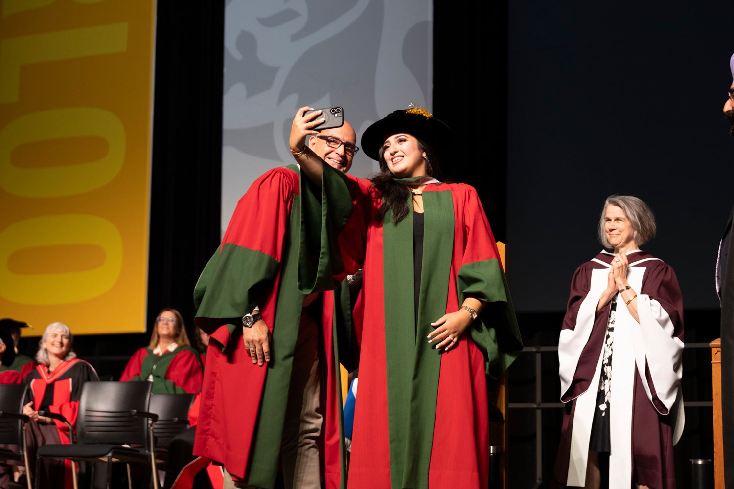 A young woman in academic regalia takes a selfie with a faculty member on stage at convocation