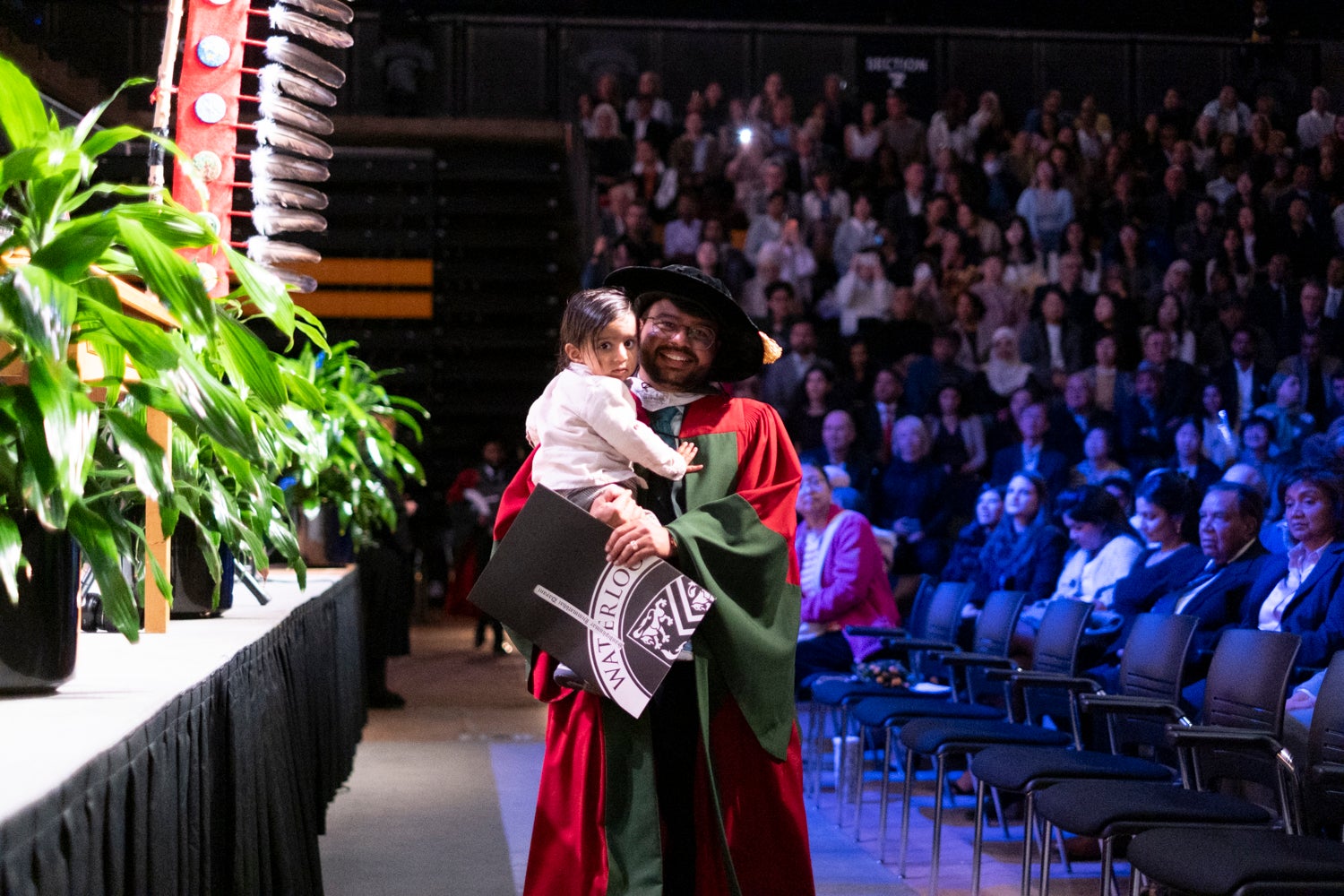 A young man with his child in his arms holds his diploma as he walks to his seat after crossing the stage