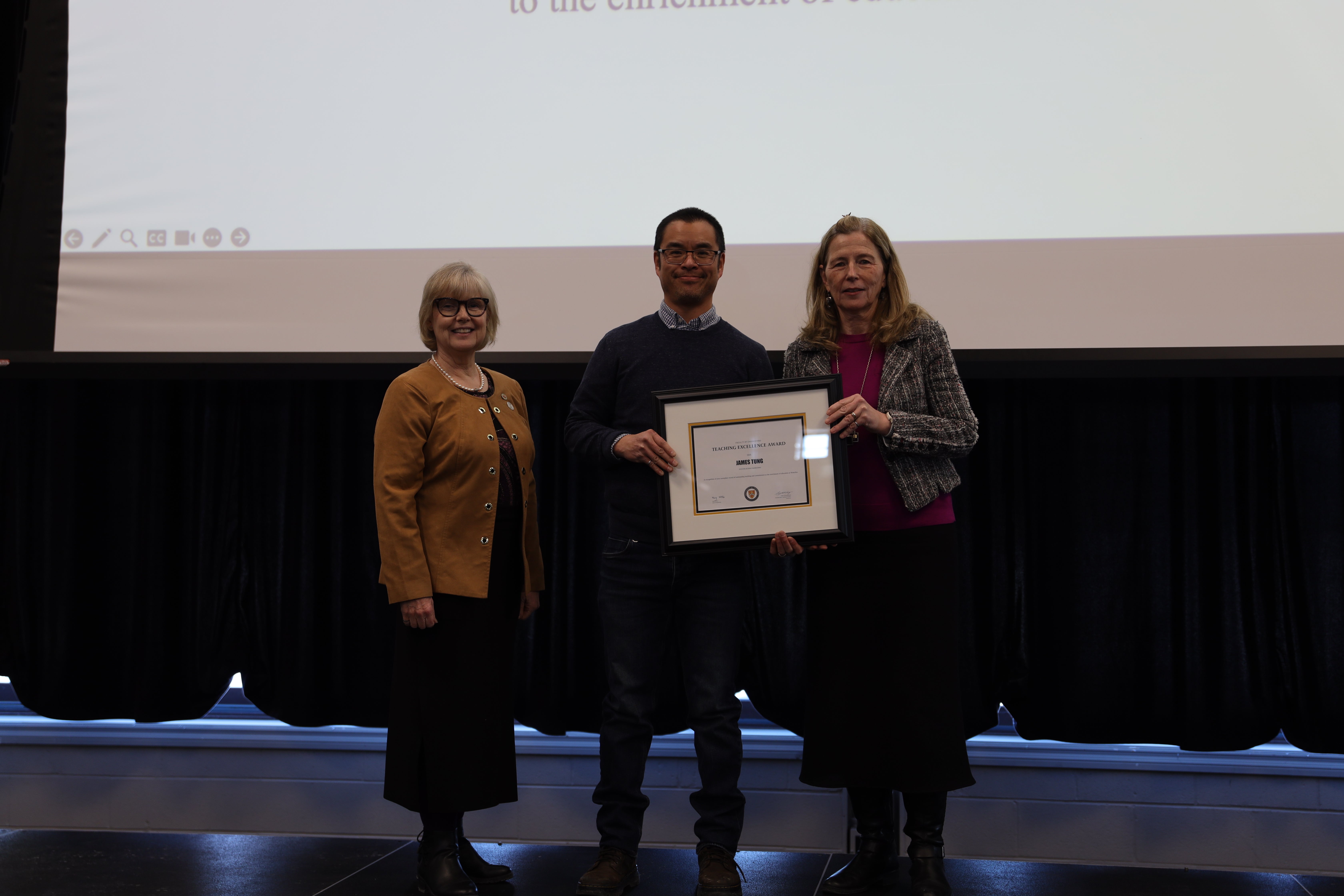 A woman, a man and another woman pose on stage; the man holds a certificate.
