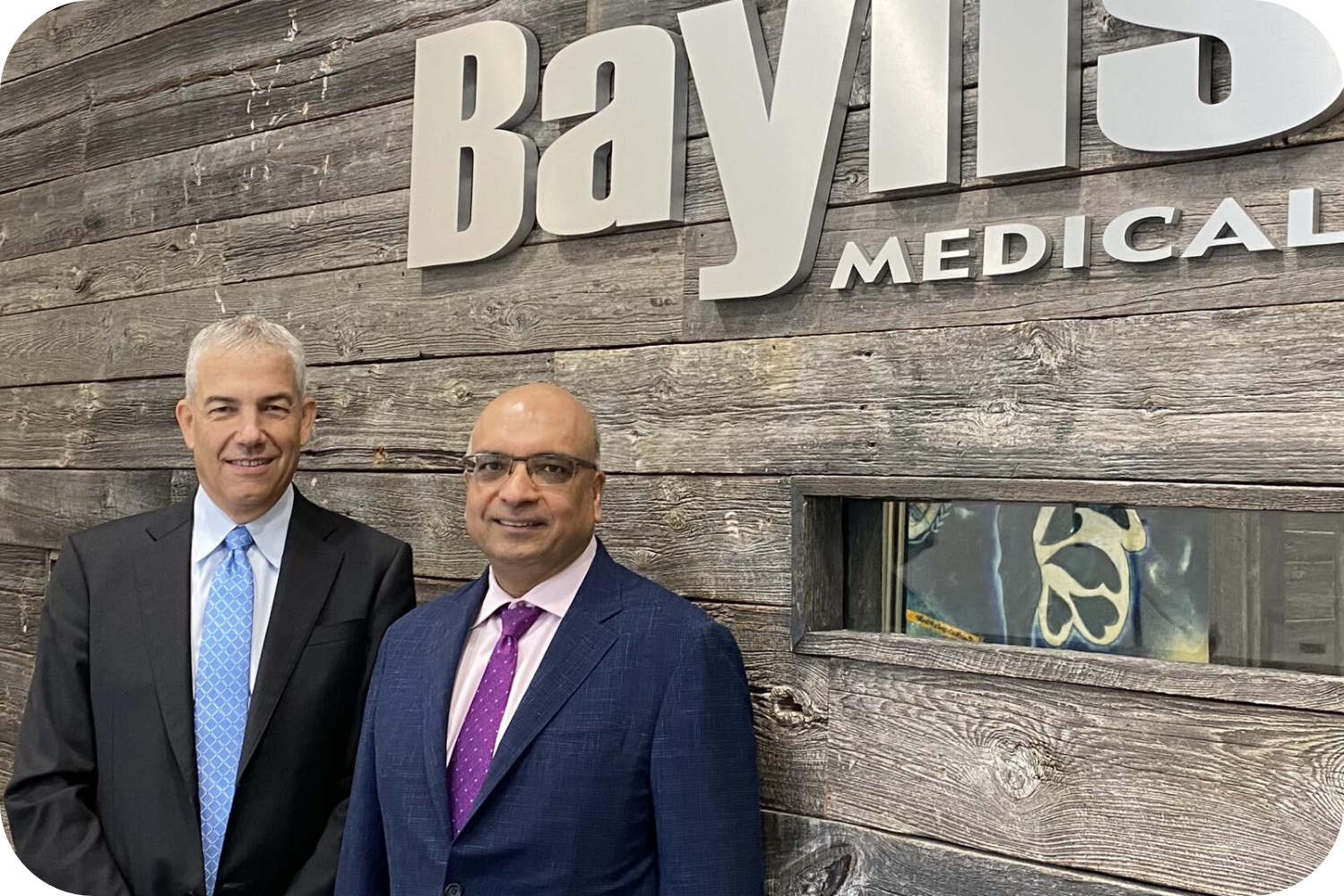Kris Shah and Frank Baylis posing in front of Baylis Medical Logo