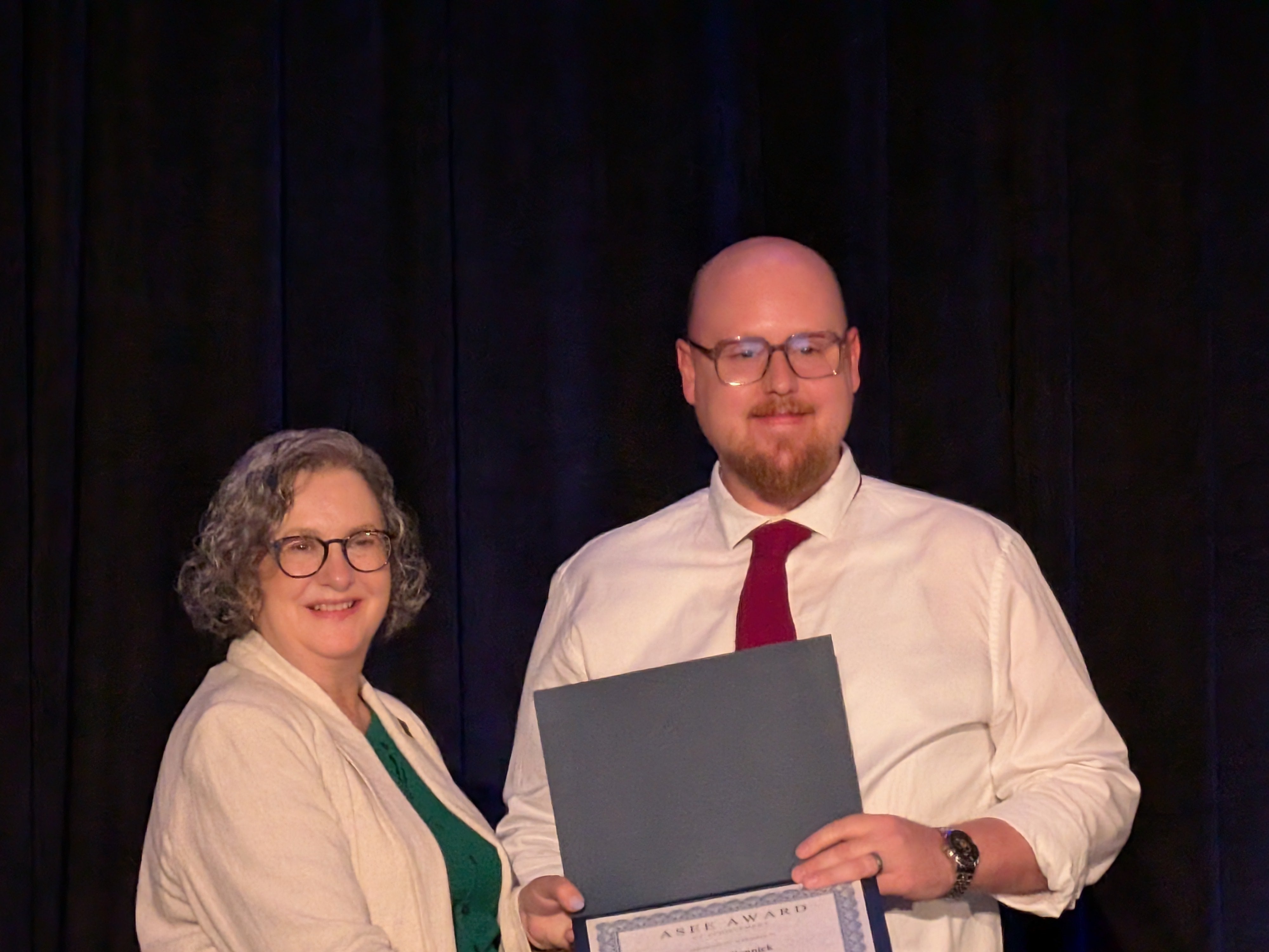 A man poses with a certificate alongside a woman with gray hair in front of a black background