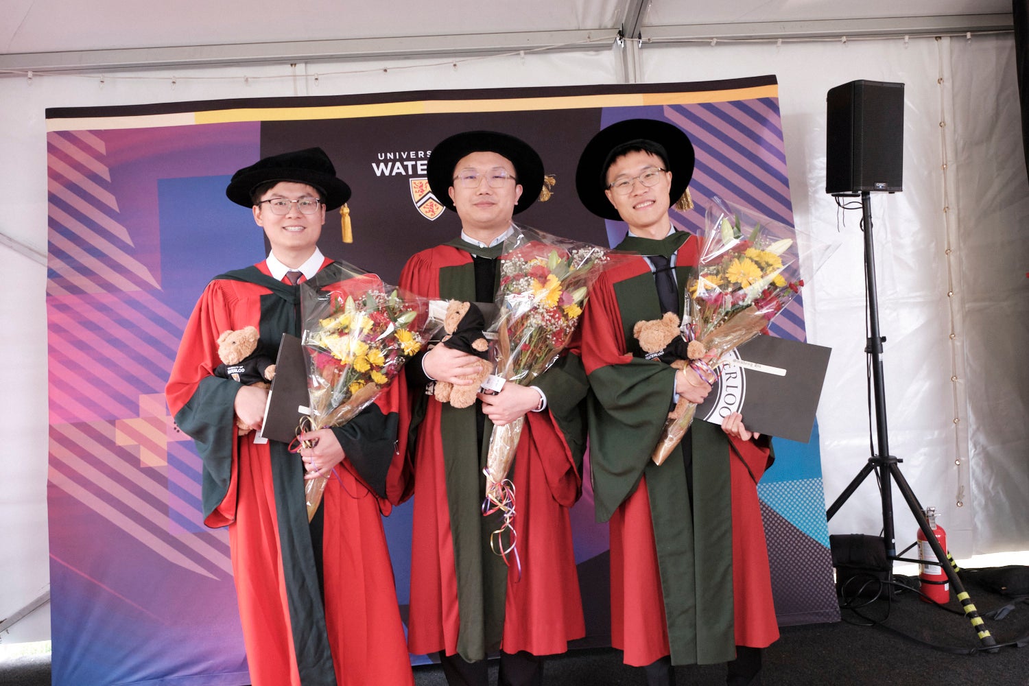 Three PhD graduates pose in front of a Waterloo Alumni sign with flowers and their certificates.