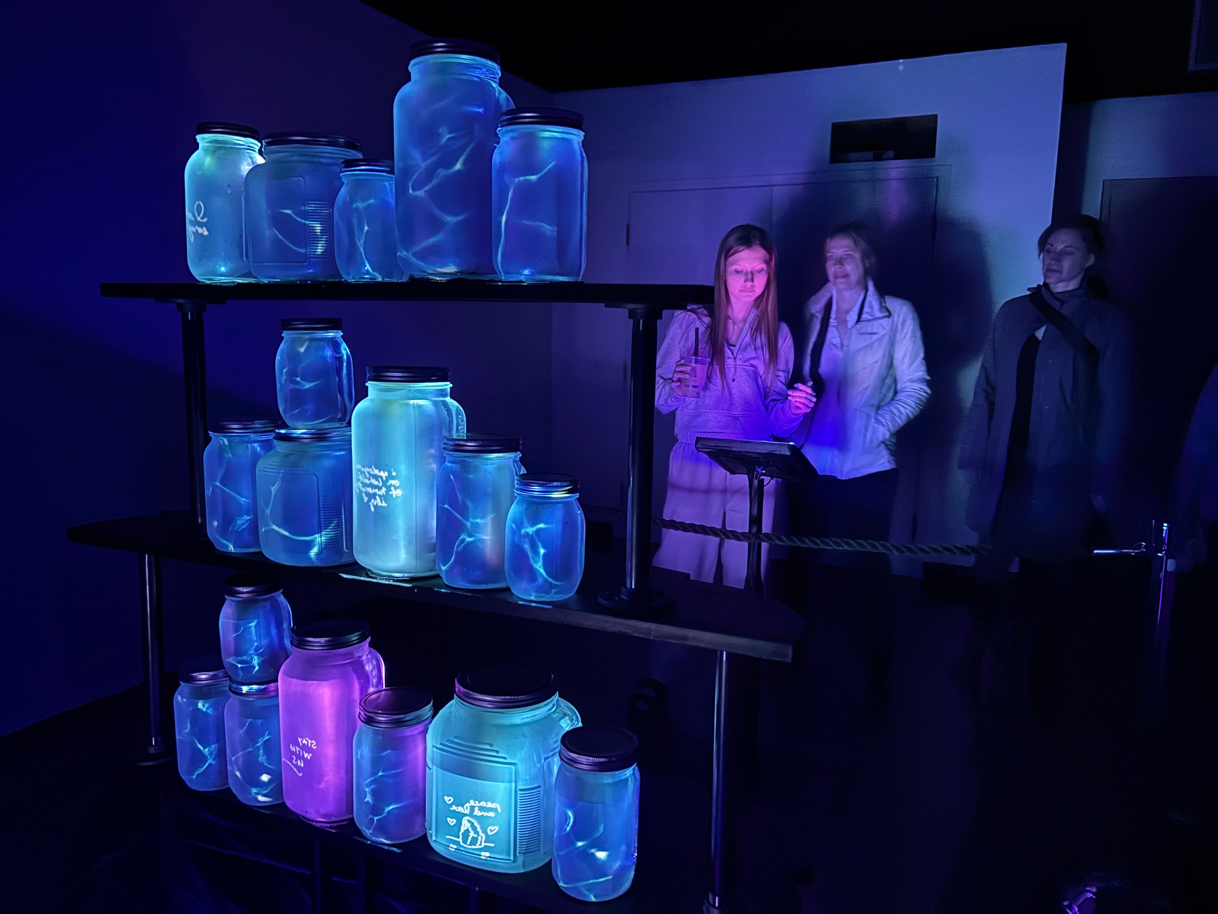 Three women look at glowing mason jars in the dark as part of an art exhibit.