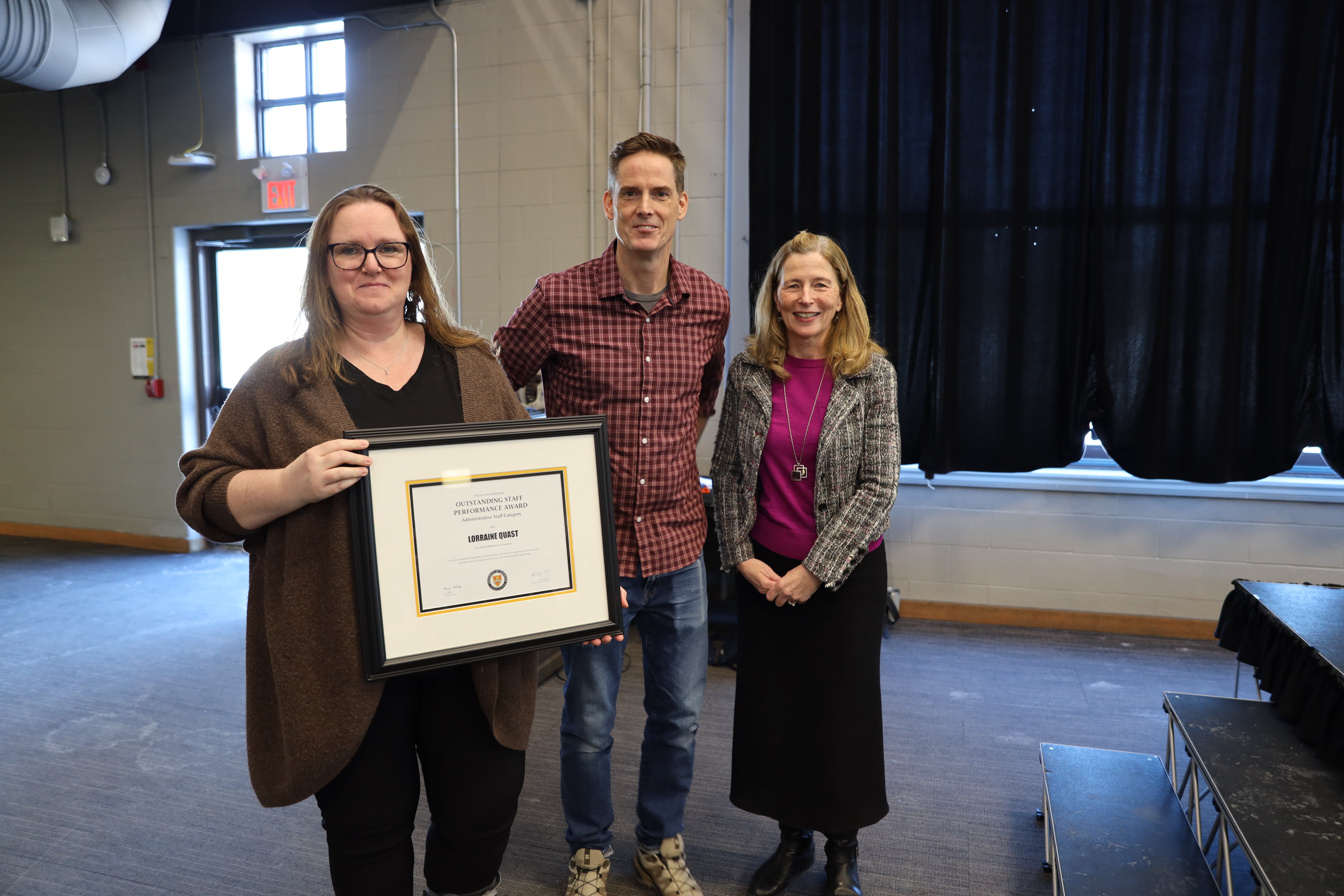 A woman, a man and another woman pose beside the stage with a certificate.