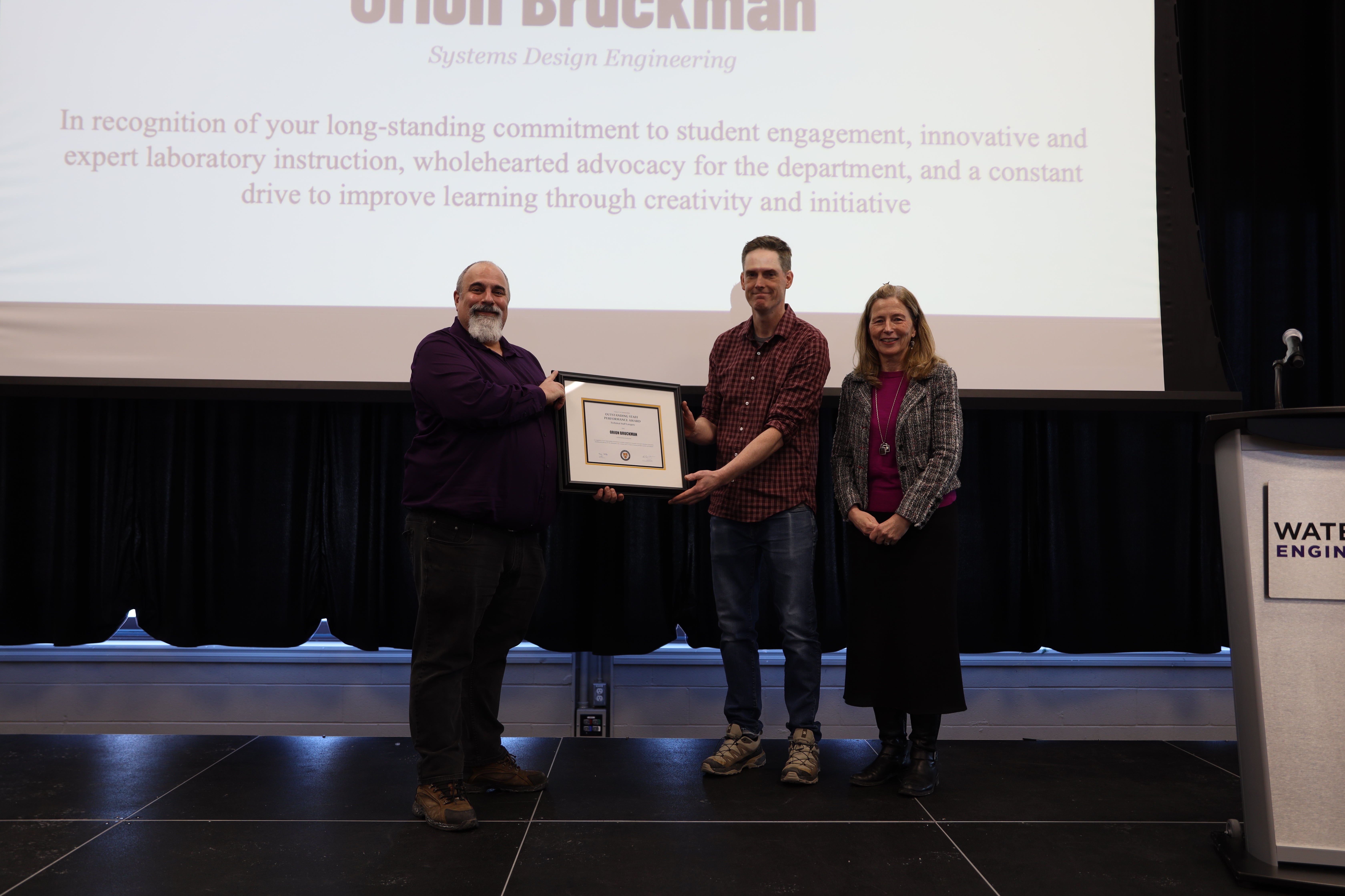Two men and one woman pose on stage with a certificate