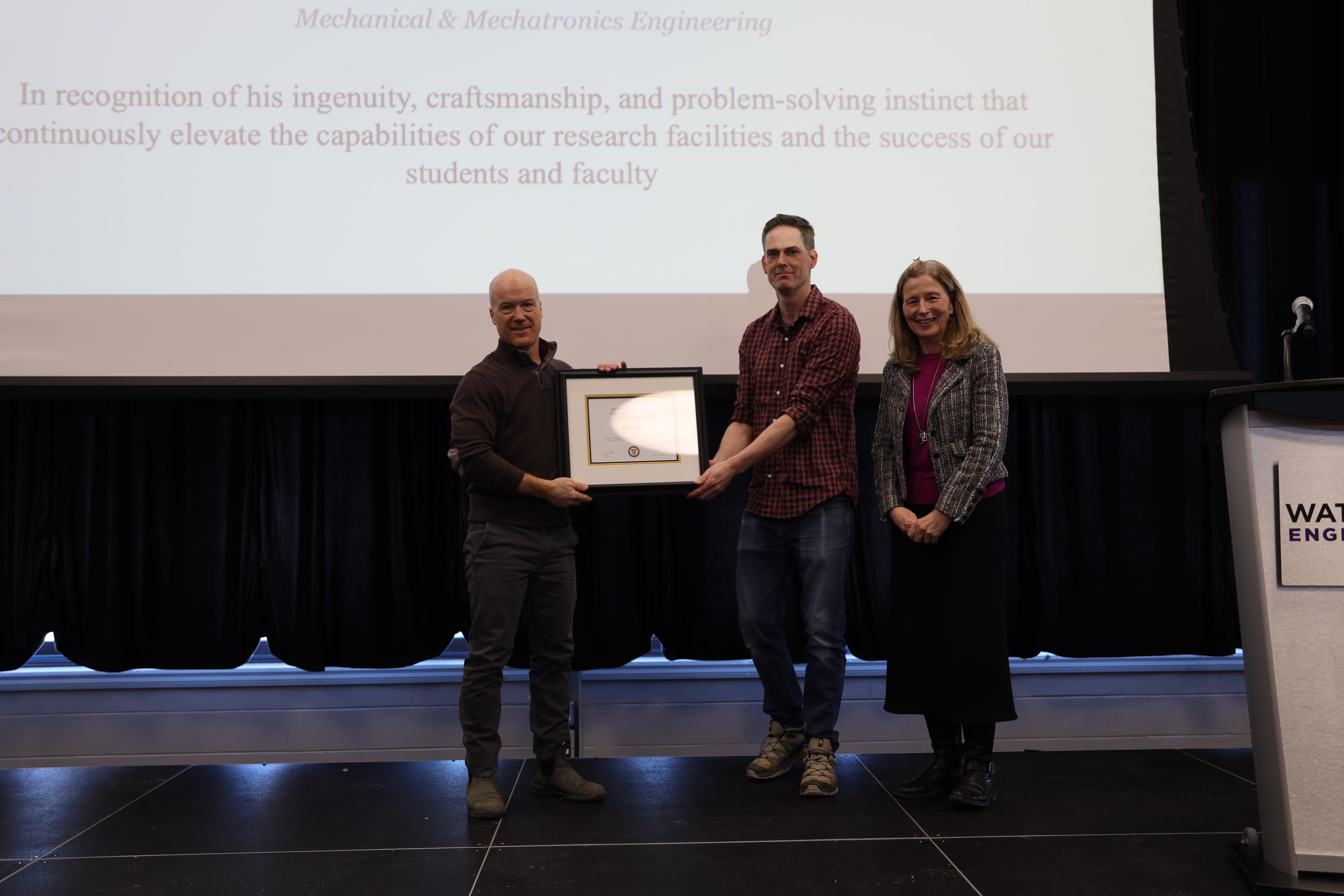 Two men and one woman pose on stage with a certificate.