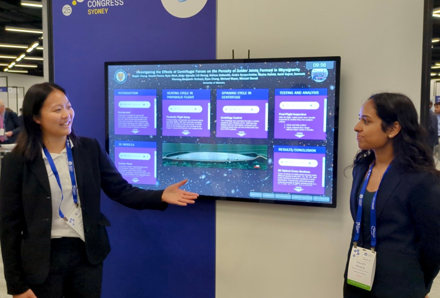 Two women stand in front of a digital screen, pointing to their presentation at a conference.