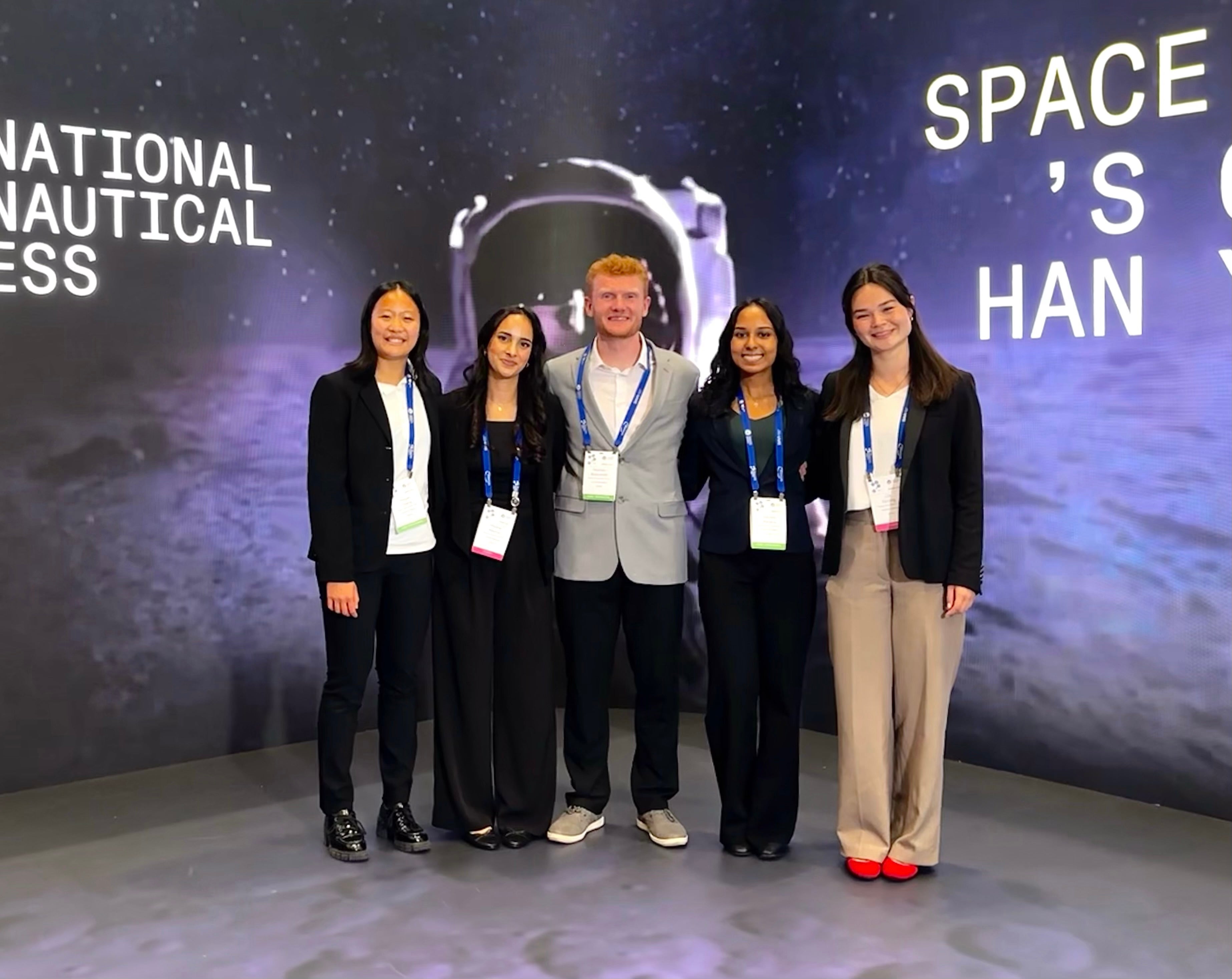 Four young women and one young man pose in front of a screen with an astronaut.