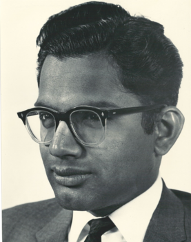 A black and white photo of a man smiling at the camera; he is wearing glasses and has dark hair