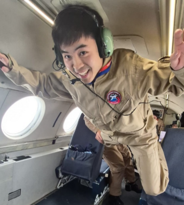 A young man floats in a plane, dressed in brown coveralls.