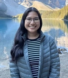 A young woman with glasses poses in front of a pond and mountain in the background.