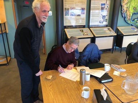 Simon Courtenay signing a document at a table with Dick Bourgeois-Doyle standing nearby.