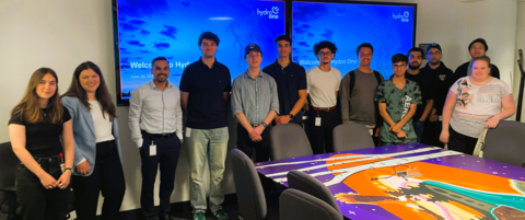a group of 13 people stand in a row facing the camera, in front of a meeting table with the Hydro One logo behind on a TV screen