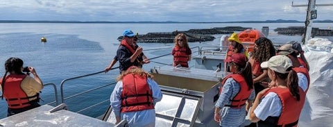 10 students visit a salmon farm in Passamaquoddy Bay, New Brunswick. They are on a boat looking at an instructor wih the Bay in the background. 