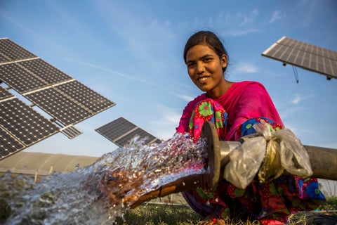 A farm worker squating next to a hose outlet with water coming out with several solar panels in the background.