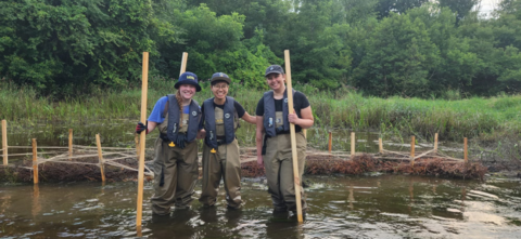 three people in hipwaders stand in a stream holding sticks