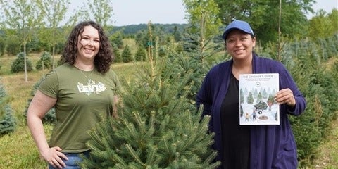 Alison Clarke and Kelsey Leonard standing among rows of small evergreens. Kelsey is holding a Growers Guide book.