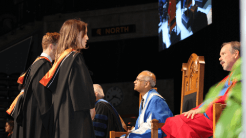 2 students in black gowns speak with professors in chairs on the graduation stage