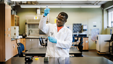 Augusting Osei in a white lab coat in a lab setting using a long pipette to withdraw a sample from a tube.