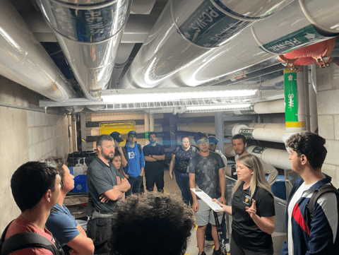 a group of people are listening to a speaker in an underground hallway that has a ceiling lined with wide pipes