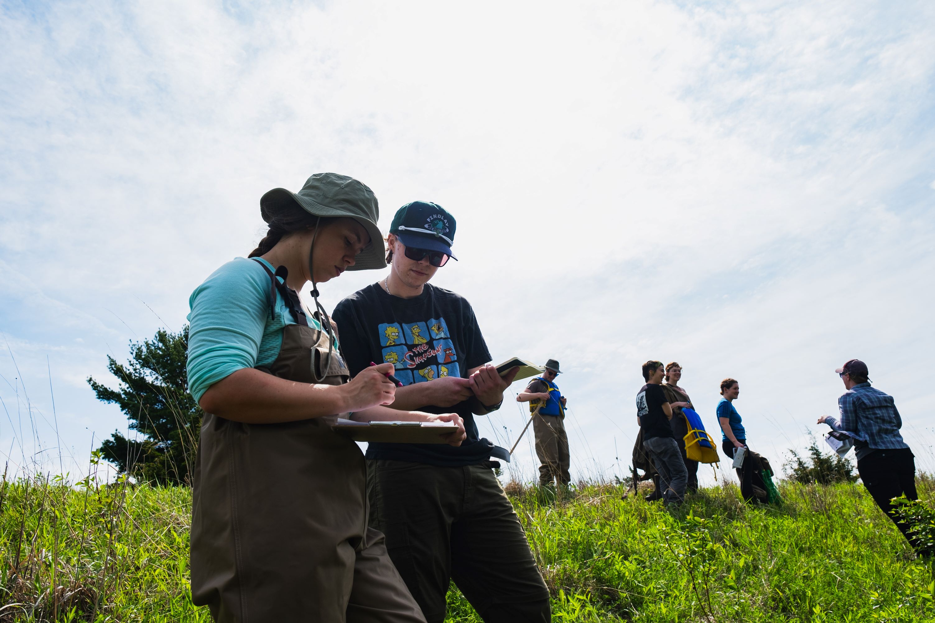 students standing on a hill compare notes. Others in the back listen to a leader.