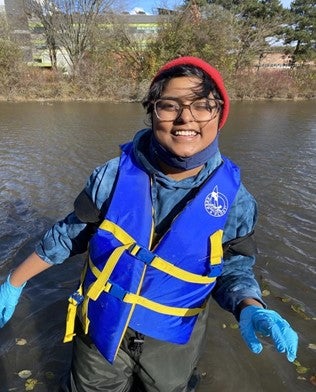 A student wearing a lifejacket, hip waders and gloves standing thigh-deep in a river looking at the camera, smiling.