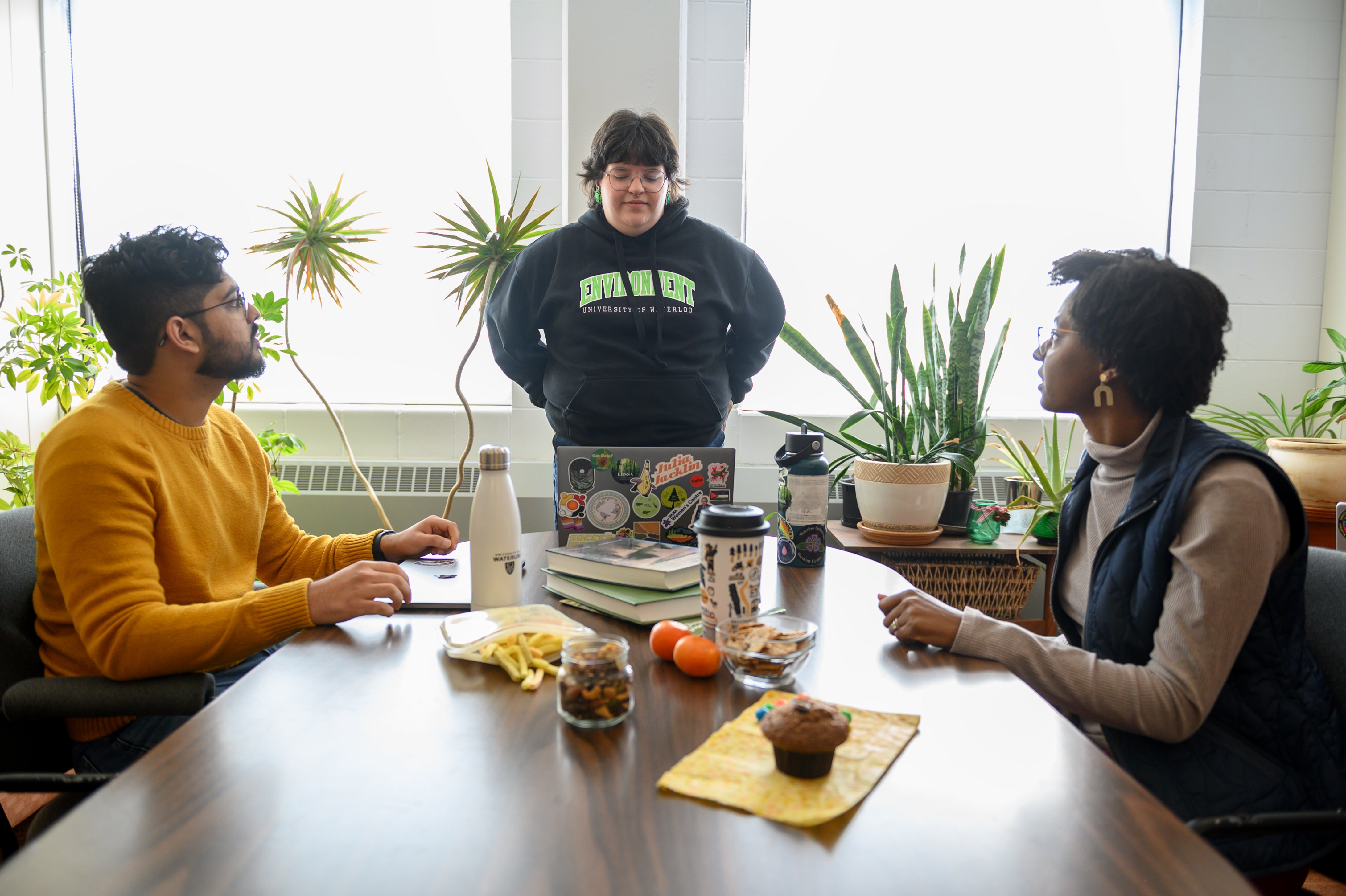 A student stands at the head of a table explaining to 2 others who sit at the table.
