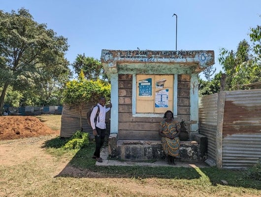 Water supply and sanitation station in Kenya with a woman sitting at the front and a man standing nearby