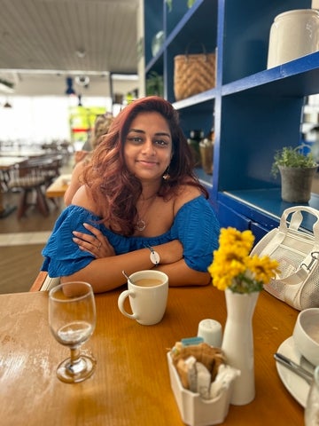 Althea (Thea) is sitting in front of a table with a water glass and mug on top. She is also wearing a blue shirt