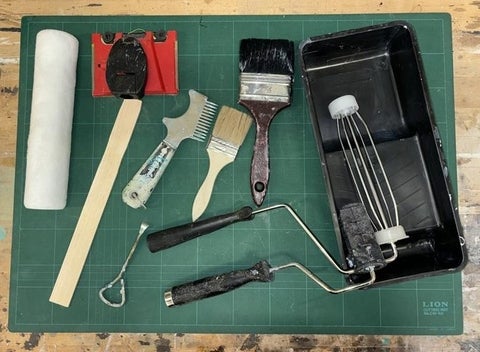 A paint roller, stir stick, paint brushes, and tray displayed on a table.