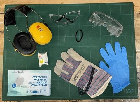 Ear muffs, a mark, ear plugs, gloves, eye protection and a hair tie displayed on a table.